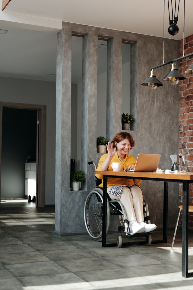 Persona con discapacidad física usuaria de silla de ruedas saluda. Esta usando una laptop.